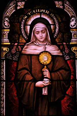 Stained glass artwork depicting Saint Clare holding a religious object, framed by an arched decorative background with rich colours.