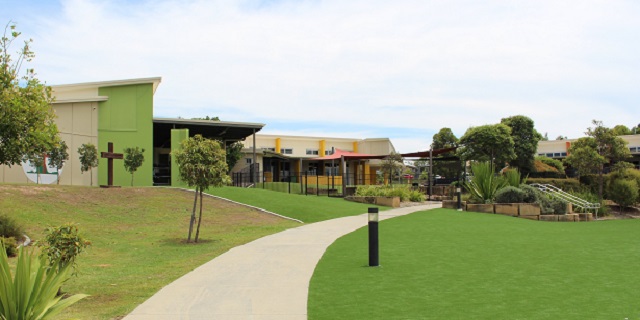 School campus with a winding pathway through a grassy area, leading past modern buildings, garden beds, trees, and a cross mounted on a green wall.