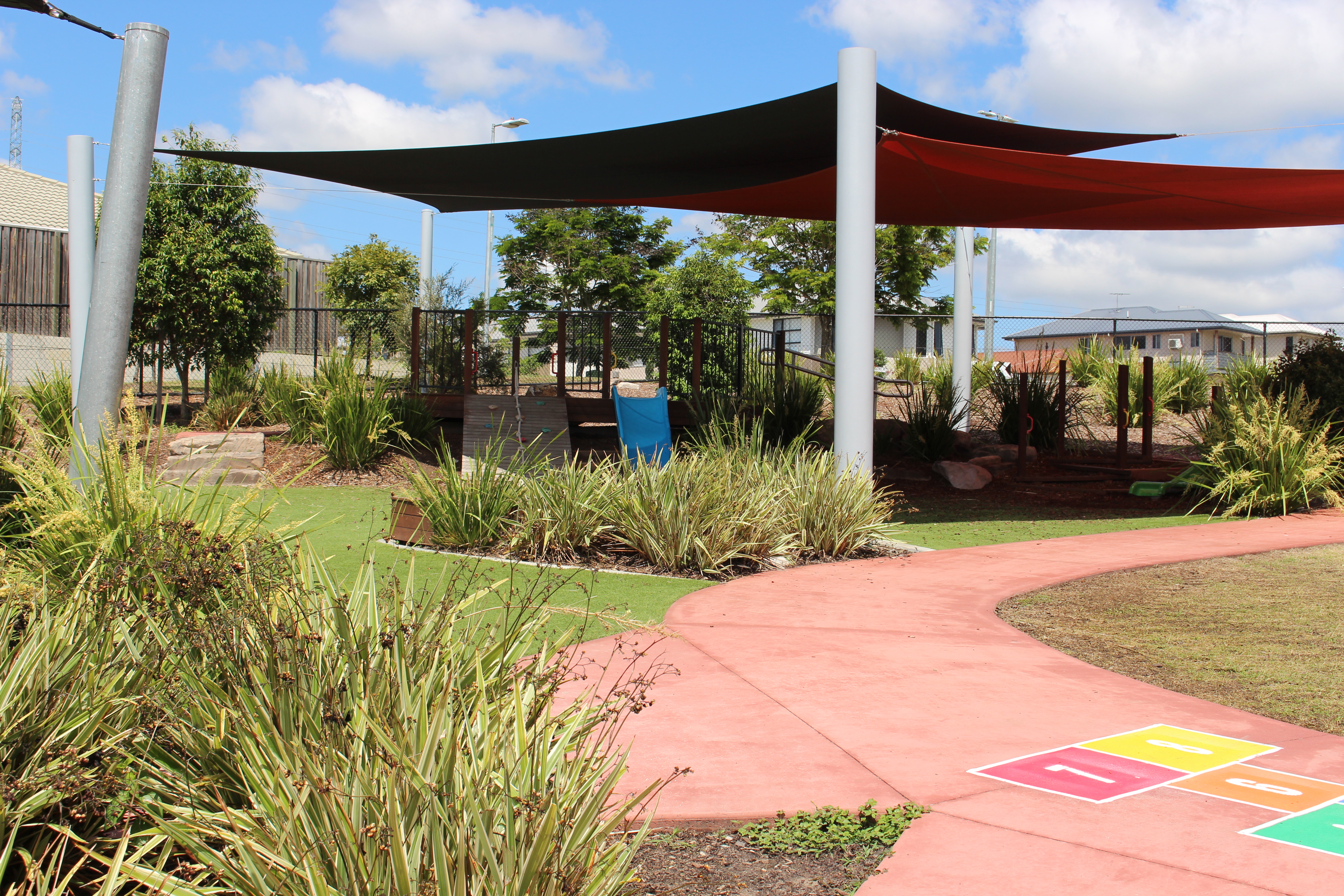 Landscaped school playground with shade sails, garden beds, climbing and balance equipment, and a hopscotch path painted on a curved walkway.