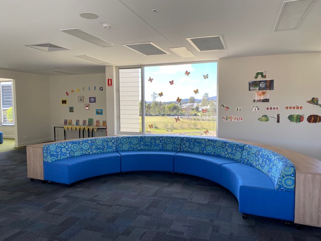 Library reading area with a curved blue seating bench, carpeted floor, wall displays, and a large window with a countryside view.