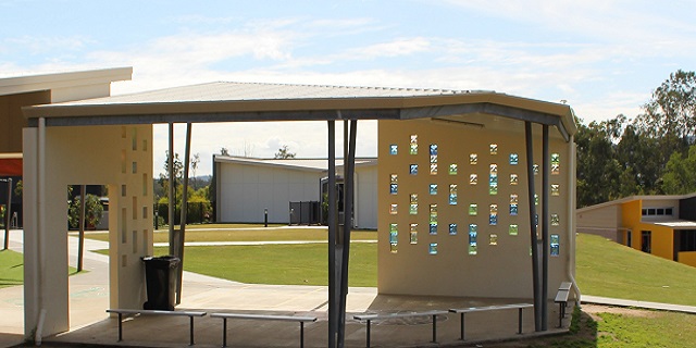 Covered outdoor seating area with benches under a shelter, featuring decorative cut‑out wall panels and a grassy area beyond.