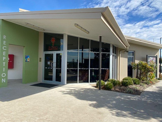 School reception entrance with glass doors, a green wall labeled “Reception,” and landscaped garden beds beside a modern building.