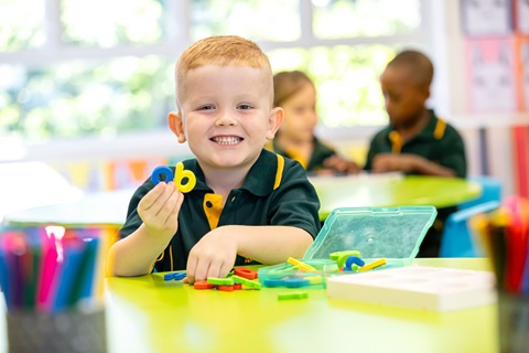 Child sitting at a classroom table playing with colourful linking shapes, with other students and classroom materials in the background.
