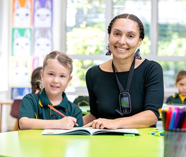 Adult and a student sitting at a classroom table, working together on a workbook, with other students and artwork visible in the background.