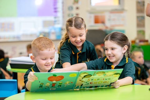 Three children in school uniforms reading a large picture book together at a classroom table.