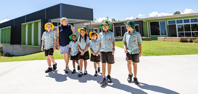Group of students in school uniforms walking with an adult along a paved path outside modern school buildings.