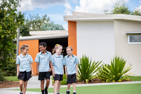 Four students in school uniforms walking together outdoors past a modern school building and garden plants.