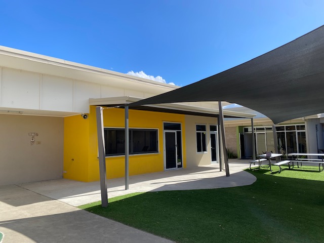 Shaded outdoor area of a school with a yellow building, covered walkway, artificial grass, and picnic tables beneath shade sails.