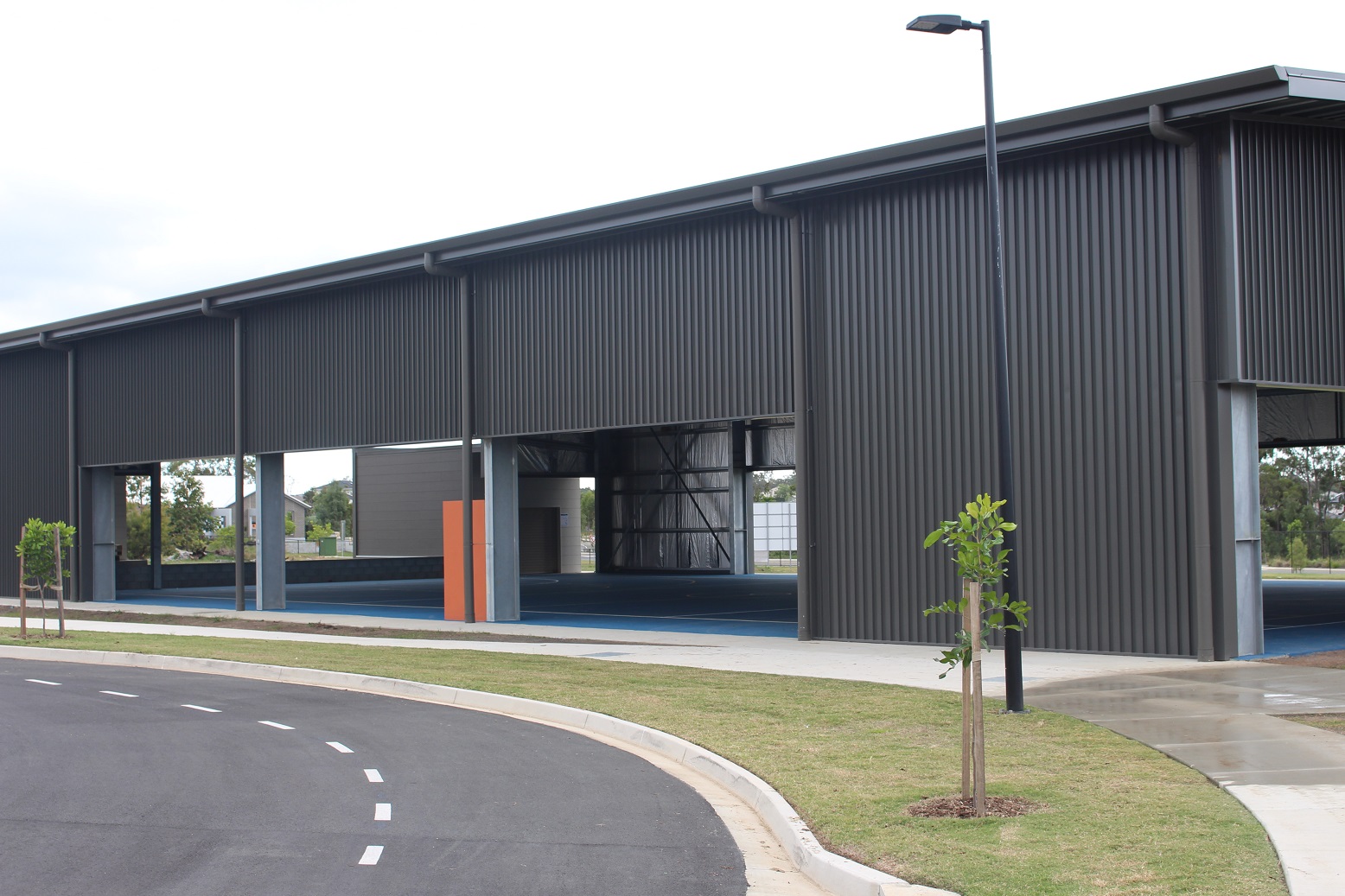 Large covered outdoor sports court with open sides, supported by steel columns, beside a roadway and grassed area on a school campus.