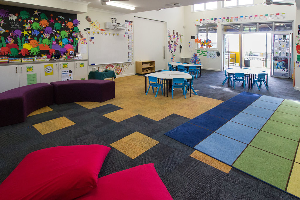 Early learning classroom with colourful wall displays, small tables and chairs, cushioned seating, and carpeted learning areas, opening onto an outdoor space through glass doors.
