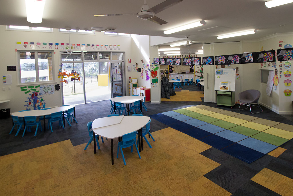 Early learning classroom with small tables and chairs, play areas, student artwork on walls, and a reading space, opening onto an outdoor play area through glass doors.